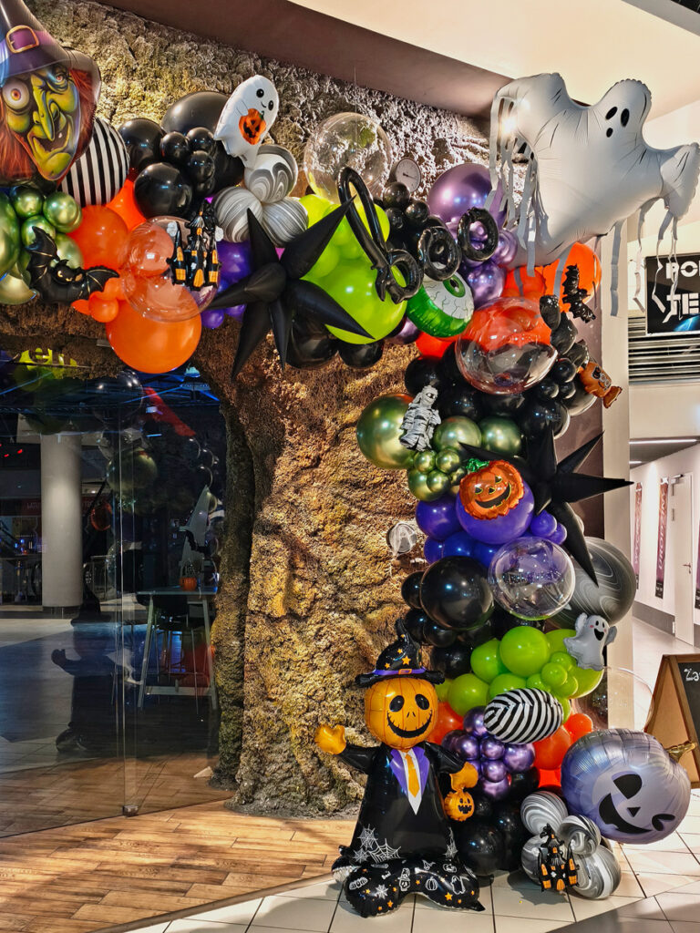 Dekoracja halloweenowa z balonów z dużym białym duchem, dyniami, upiornym okiem i czarnymi gwiazdami przy wejściu do lokalu.