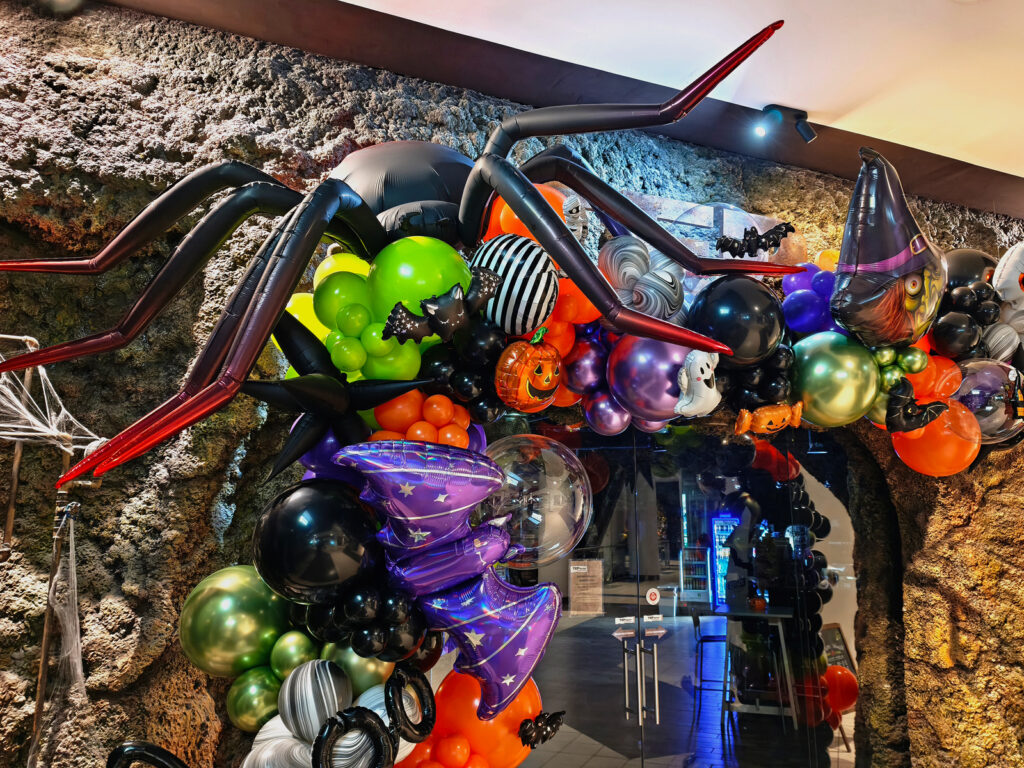 Gigantyczny czarny pająk nad organiczną girlandą balonową na Halloween, ozdobioną dyniami, nietoperzami i czarownicą.